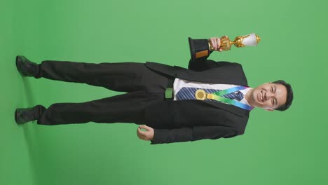 슈트와 타이를 입은 아시아 사업가, 황금 메달을 입고 손에 황금 트로피를 들고, 스튜디오의 녹색 화면 배경에서 카메라에 미소 짓는 첫 번째 우승자입니다.