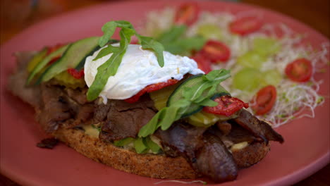 Slow-motion-close-up-of-a-beef-deli-sandwich-topped-with-arugula-mozzarella-cheese-pickles-cucumber-and-tomato-served-with-salad