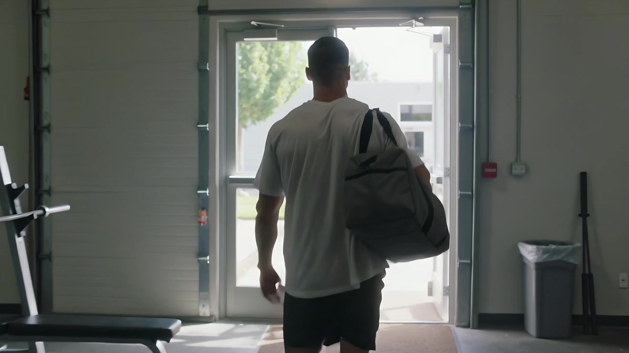 Walking man carrying gray duffel, wearing white tee dark shorts, daylight drawing toward gym exit