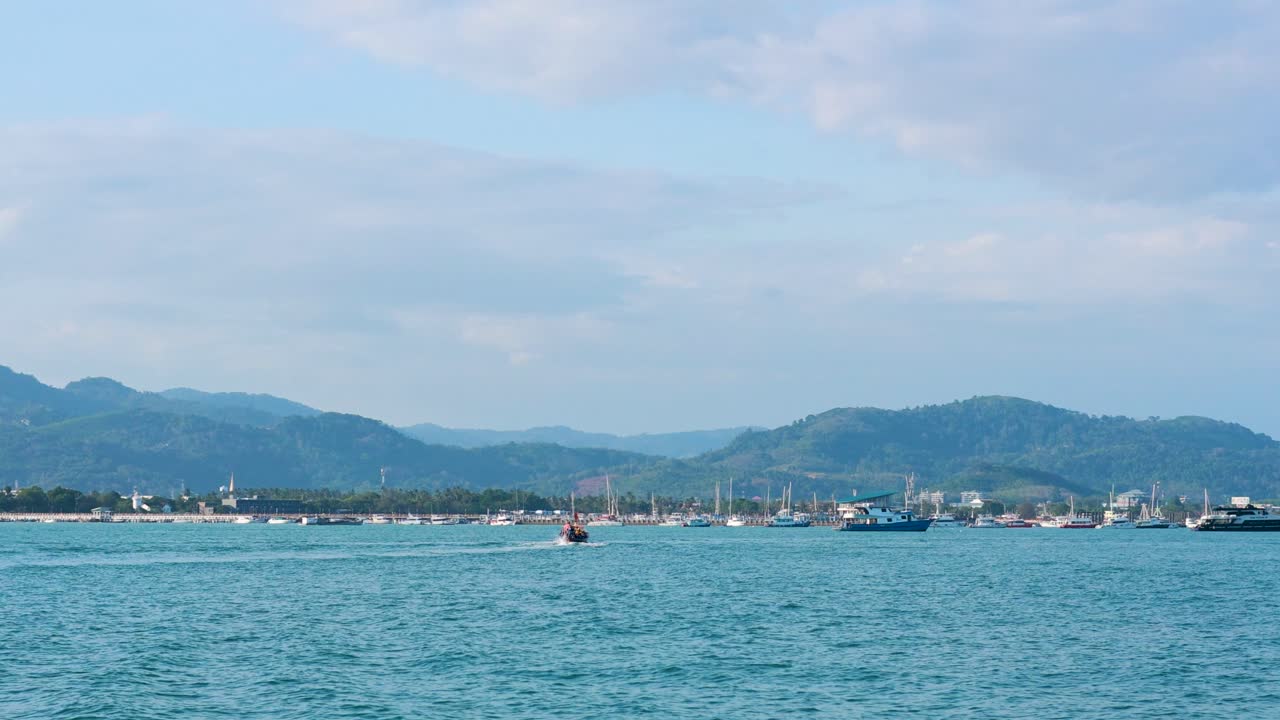 Fishing boats move across a tranquil bay in Phuket, Thailand, with mountains in the background under clear skies