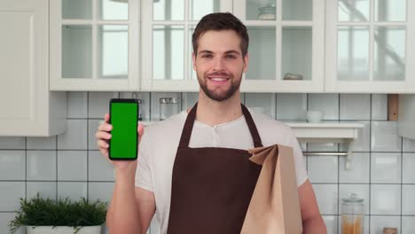 un hombre con un delantal sostiene un teléfono inteligente con una pantalla verde en la mano y sonríe.