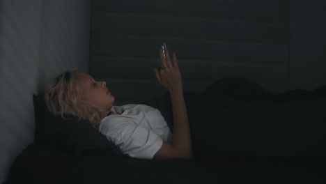 Blonde-girl-in-a-white-T-shirt-looks-at-the-phone-before-going-to-bed-in-a-gray-bed-at-night