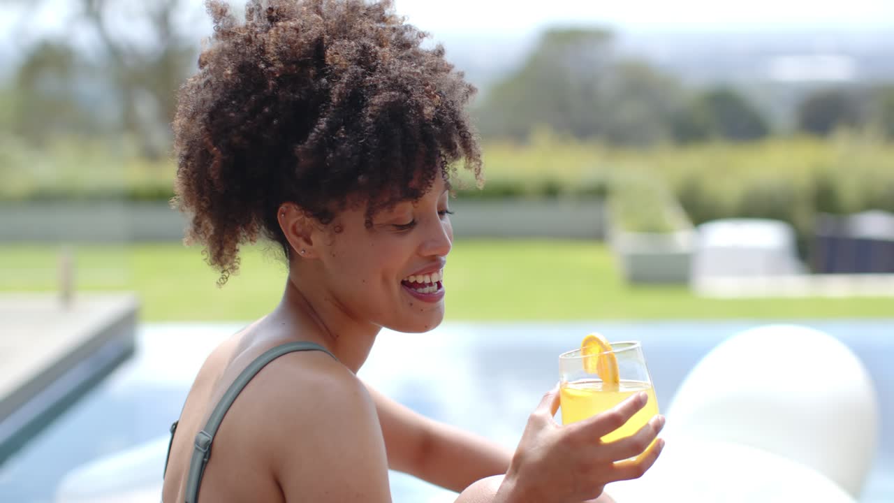 African American woman sitting at pool in swimsuit, smiling at camera, holding glass, savoring sun
