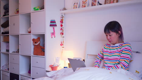 young-asian-malaysian-chinese-woman-sitting-in-bed-under-duvet-using-laptop-sideways