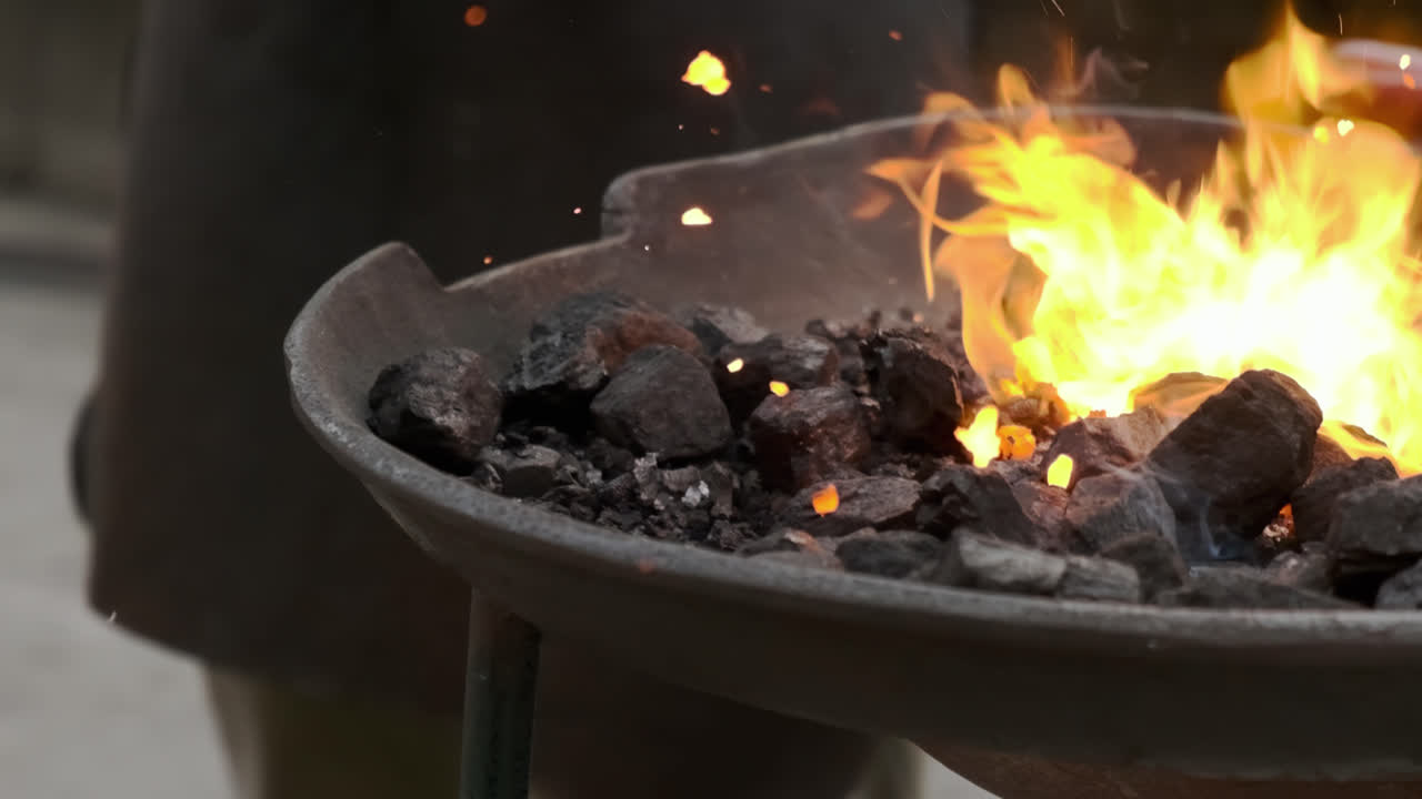 Right-to-left slow-motion pan tracking a blacksmith withdrawing a heated metal piece from a coal forge; bright flames, flying sparks and embers, close-up industrial atmosphere