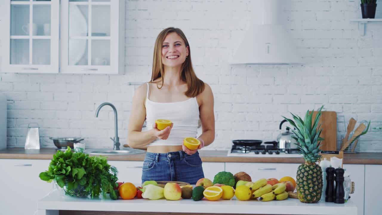 Beautiful female in the light kitchen. Smiling young woman proposing fresh orange. Healthy organic fruit and vegetables on the table.