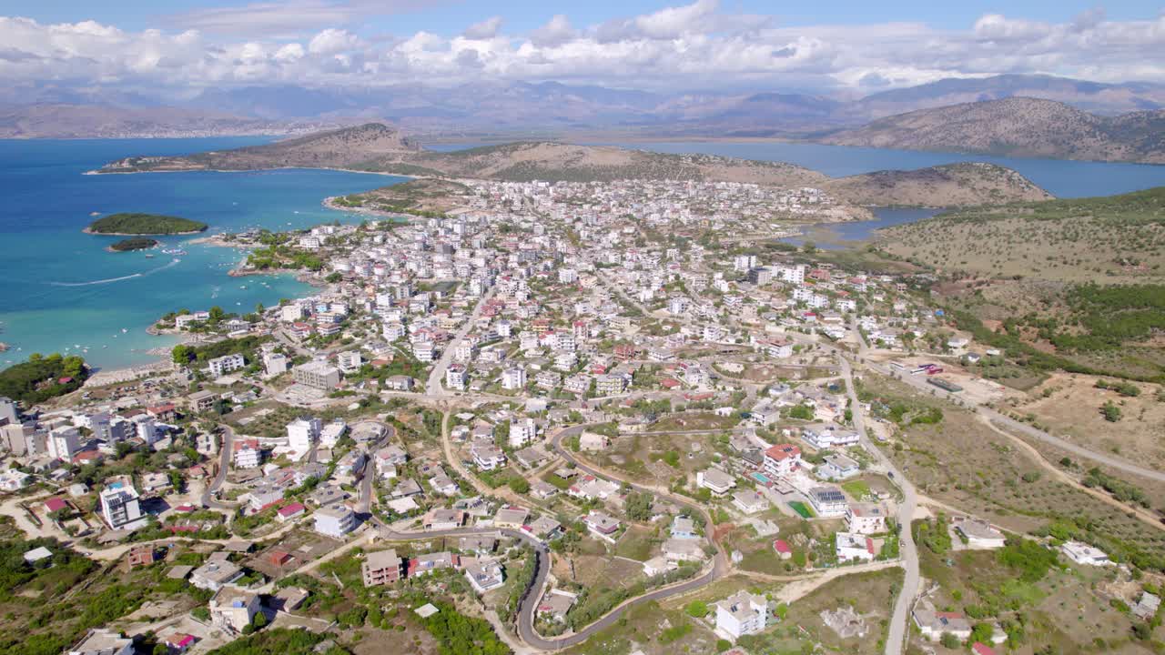 Highaltitude drone view shows a dense coastal town with winding roads, turquoise bays, small islands, and surrounding hills under a partly cloudy sky, a topdown aerial perspective