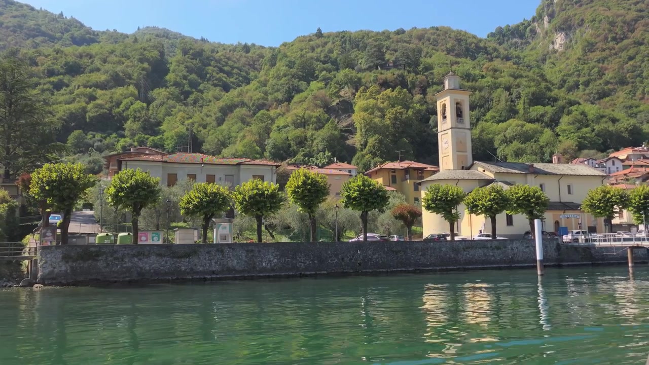Pittoresco villaggio lacustre caratterizzato da case classiche, rigogliose colline verdi, una chiesa con campanile e acque cristalline sotto una luce diurna brillante in un'inquadratura grandangolare