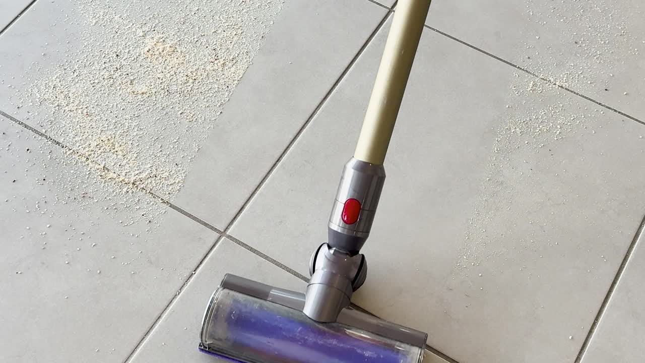A cordless vacuum cleaner removes scattered crumbs from a light-colored tiled floor. Bright natural lighting highlights the cleaning process with smooth, steady camera movement
