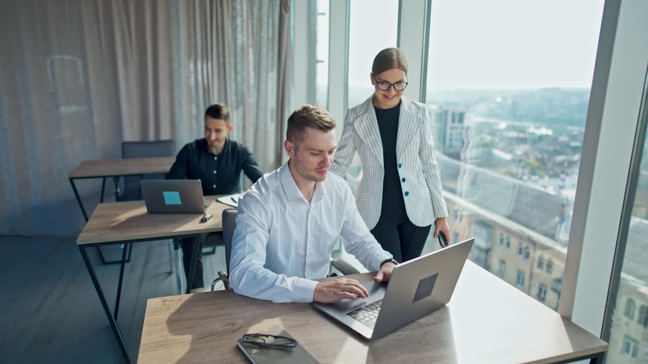 Young office team working in the light modern office. Men working at laptops and woman standing by one desk. Cityscape at backdrop in blur.