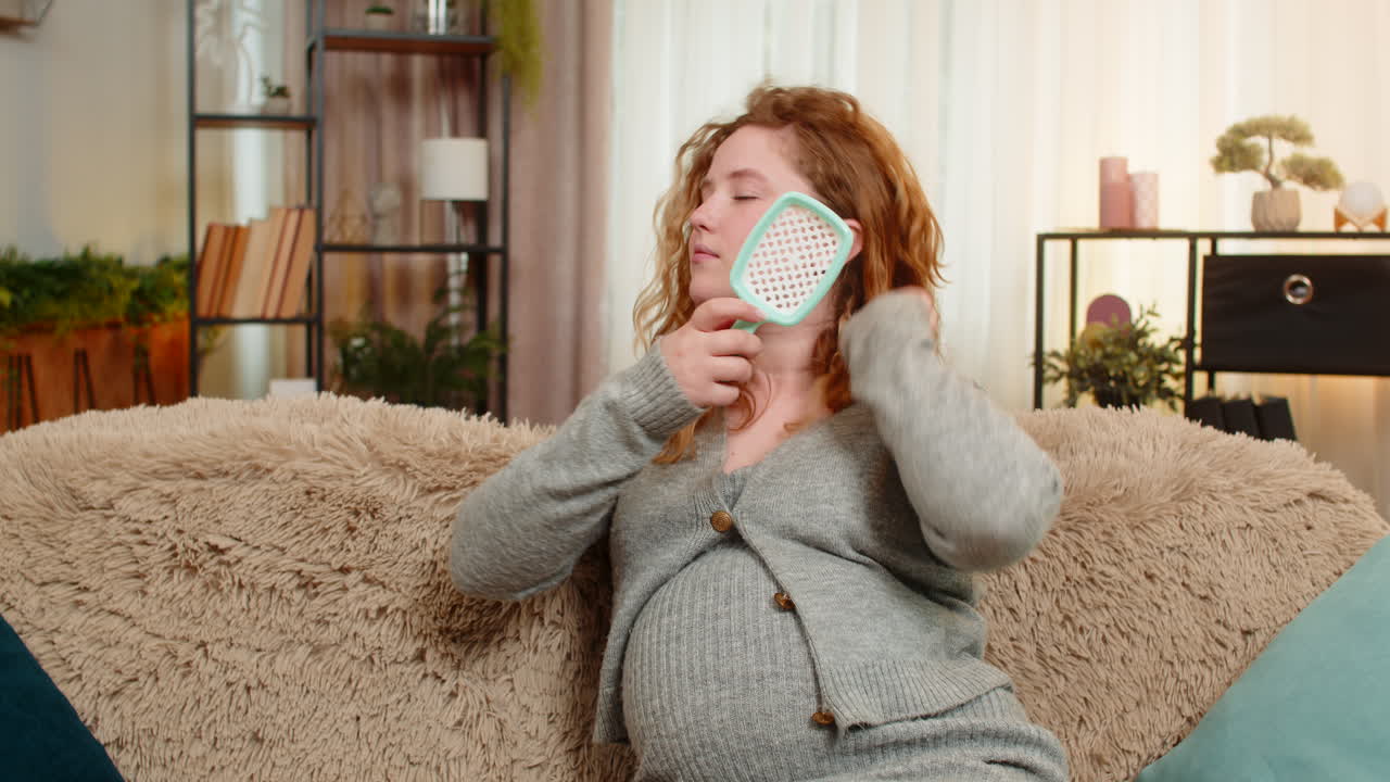 Pregnant young woman brushing hair on home sofa relaxed and calm enjoying peaceful and quiet moment