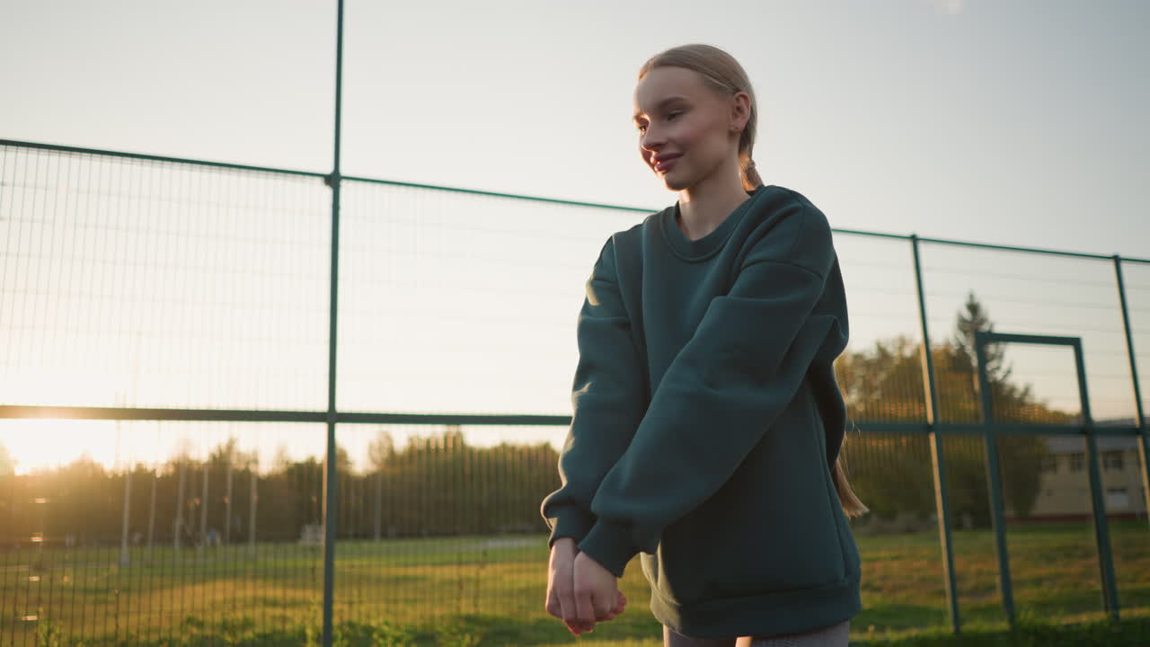 Young lady in green hoodie playing volleyball with another player in background, with sunlight glow effect in distance, set against open field and a building in the background during golden hour