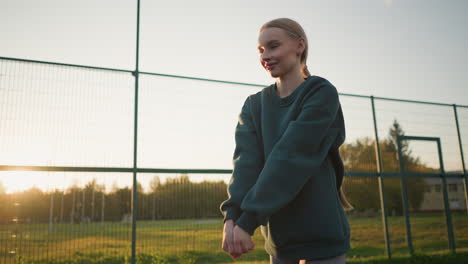 young lady in green hoodie playing volleyball with another player in background, with sunlight glow effect in distance, set against open field and a building in the background during golden hour