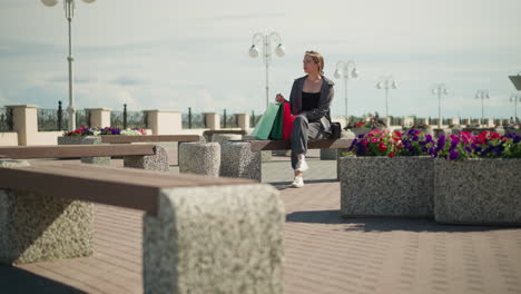 white lady seated on an outdoor bench with colorful shopping bags next to her, looking off into the distance, flowers nearby flutter in the wind while light poles can be seen in the background