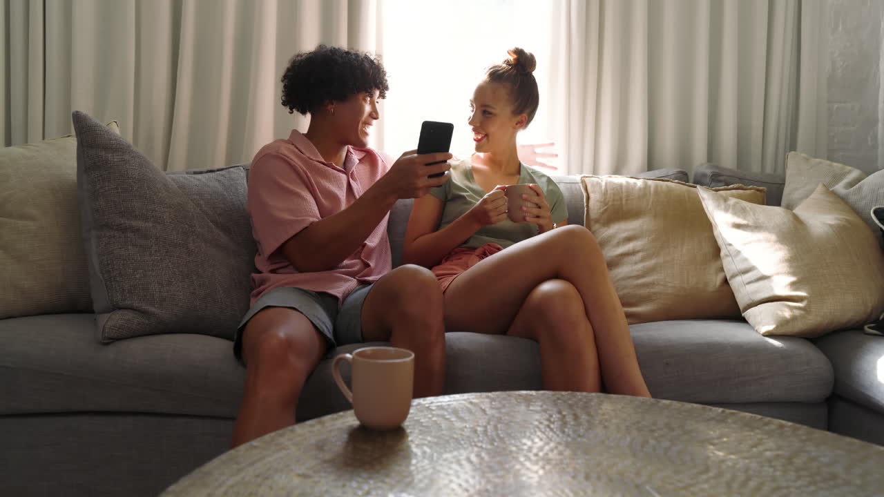 Coppia multietnica che si rilassa sul divano, sorridendo e condividendo uno smartphone, godendosi un pomeriggio accogliente a casa.