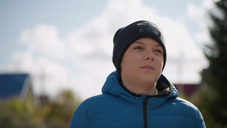 close-up of young boy in blue jacket and black beanie strolling in residential area, looking thoughtful, with blurred houses and autumn trees in the background