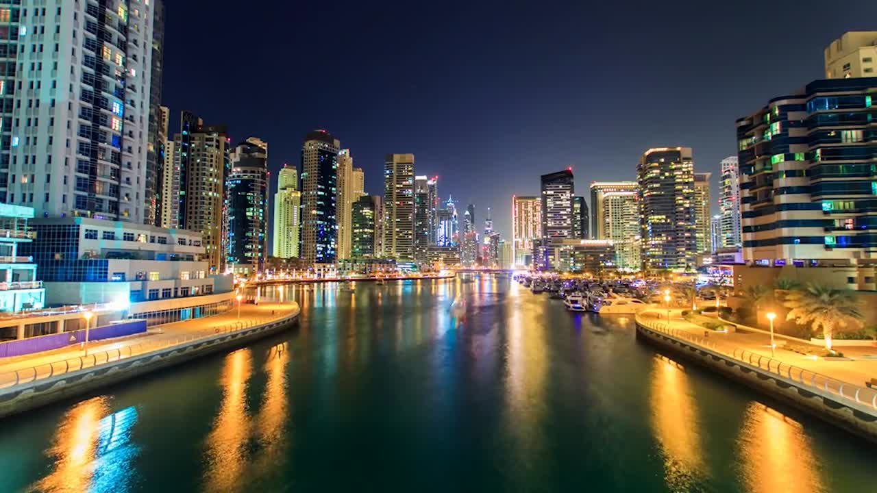 Séquence en accéléré de la marina de Dubaï la nuit, présentant des gratte-ciel illuminés et des reflets colorés sur l'eau. Un bateau éclairé navigue sur le cours d'eau central, entouré de yachts amarrés
