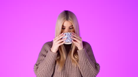 portrait of pretty blonde woman enjoying drinking from coffee mug, looking at camera against pink background, studio shot