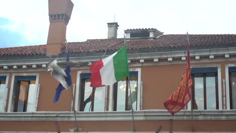 An-Italian-flag,-European-flag,-and-the-flag-of-Venice-waving-on-the-facade-of-a-building-on-a-sunny-day-while-a-bird-flies-by-in-slow-motion