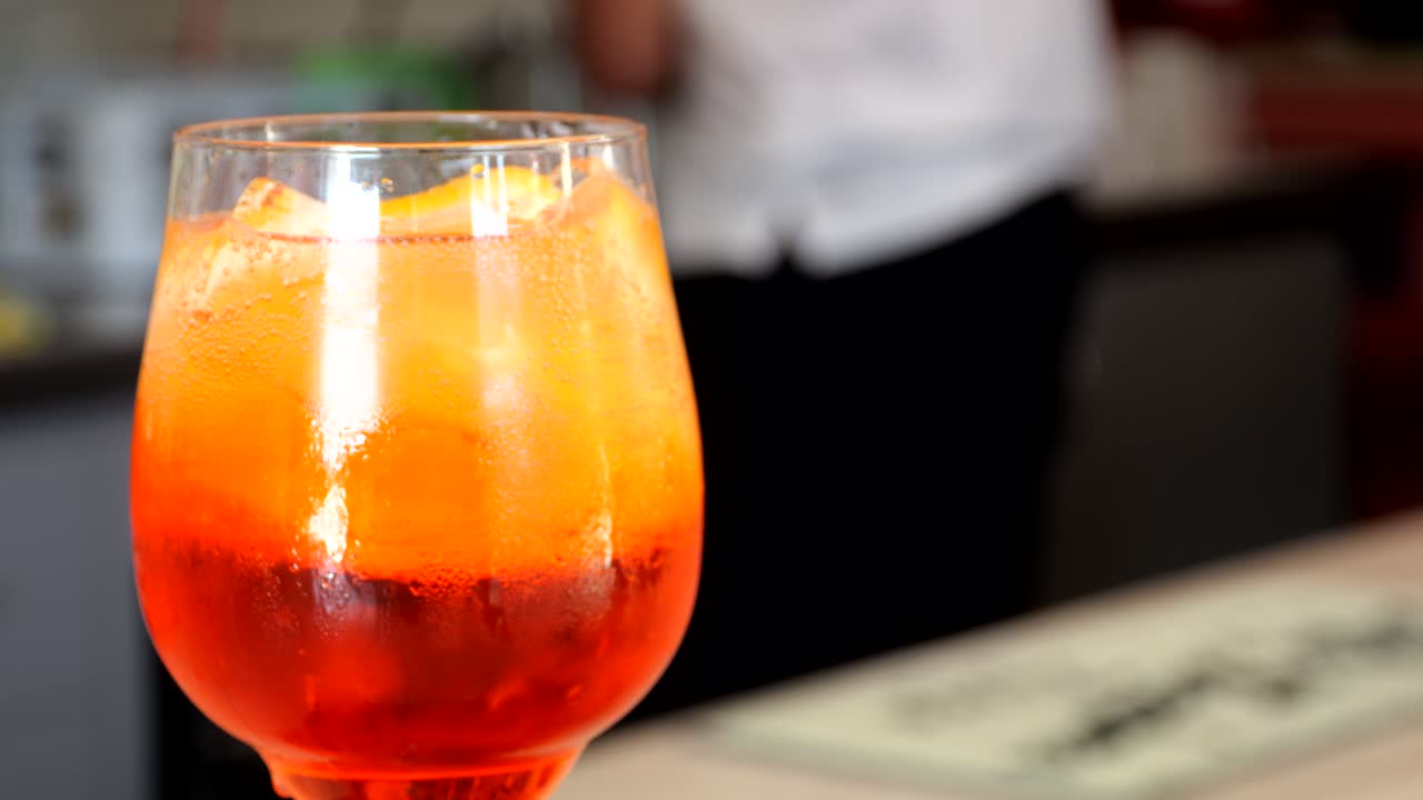 Refreshing Aperol Spritz served over ice with orange slices in a stemmed glass, resting on a bar counter with blurred bartender in the background, captured by close-up shot