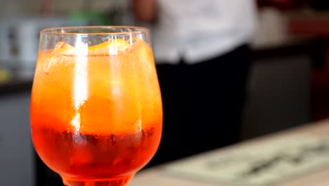 refreshing aperol spritz served over ice with orange slices in a stemmed glass, resting on a bar counter with blurred bartender in the background, captured by close-up shot