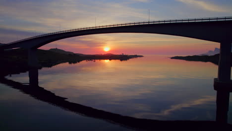 aerial forwarding drone shot, flying under fredvang bridge during midnight sun reflecting on water, lofoten