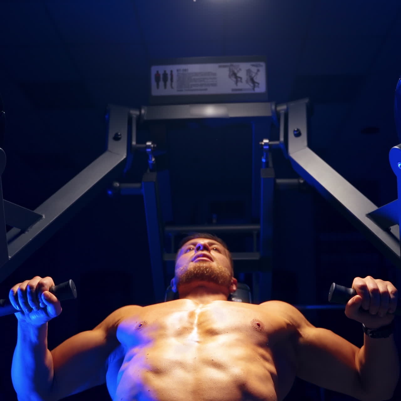 Handsome young man doing bench press workout in gym. Blue light filter. Strong abs. Heavy lift. Video from below.