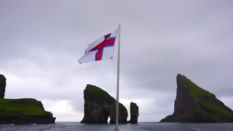 faroese flag fluttering in the wind with drangarnir sea stacks and tindholmur islet in background