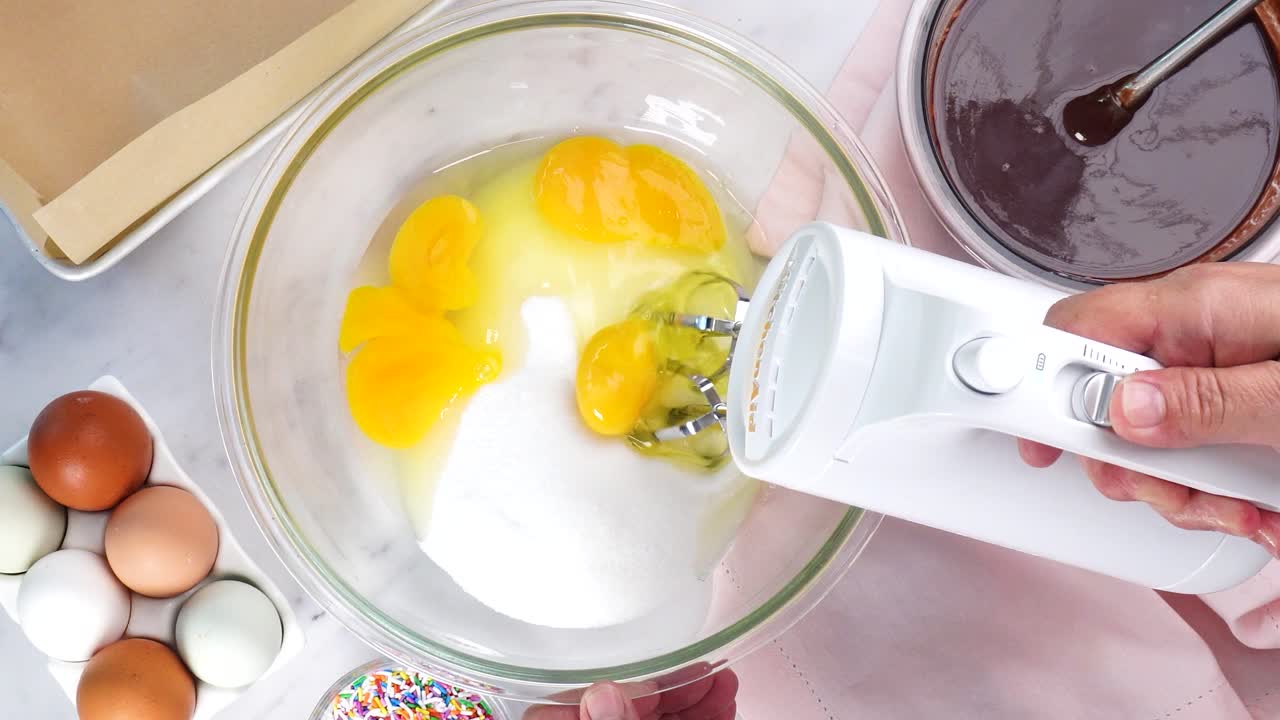 Baking chocolate brownies and using a white electric mixer to mix the batter. Making brownies on a white background surrounded by eggs, pink napkin and a baking pan. In the kitchen making a dessert