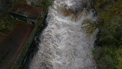 Drone-footage-directly-above-a-fast-flowing-river-and-waterfall-and-surrounded-by-old-buildings-tilts-to-follow-the-river-as-autumnal-trees-overhang-the-gorge