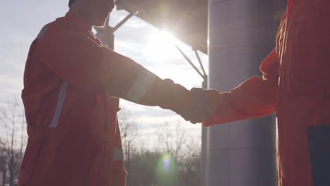 Zwei-Bauarbeiter-In-Orangefarbener-Uniform-Und-Schutzhelmen-Schütteln-Sich-Am-Bauobjekt-Die-Hand