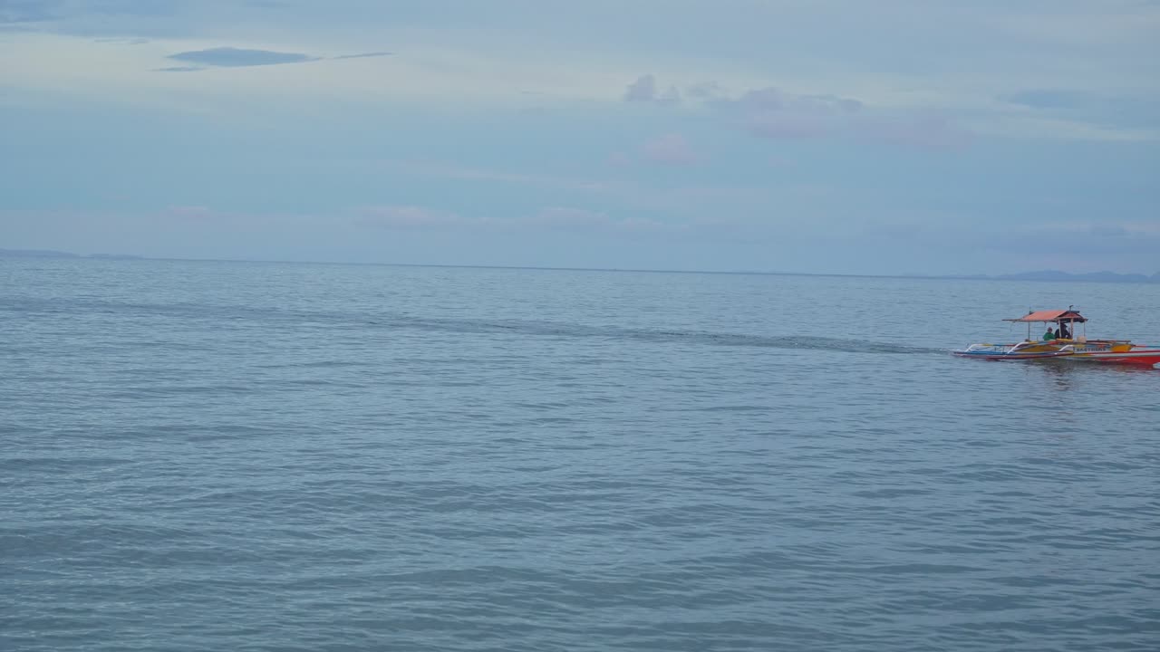 A static shot of a small red‑yellow outrigger motorboat glides across the frame getting dragged by the calm blue water of Mauban Port, Quezon Province Philippines