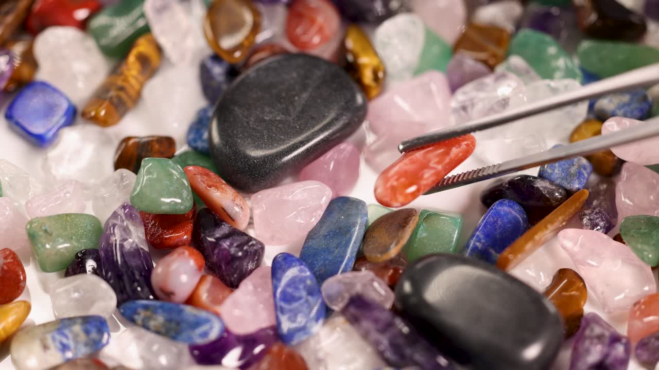 Tweezers carefully pick a red jasper stone from a colorful array of chakra stones under bright lighting
