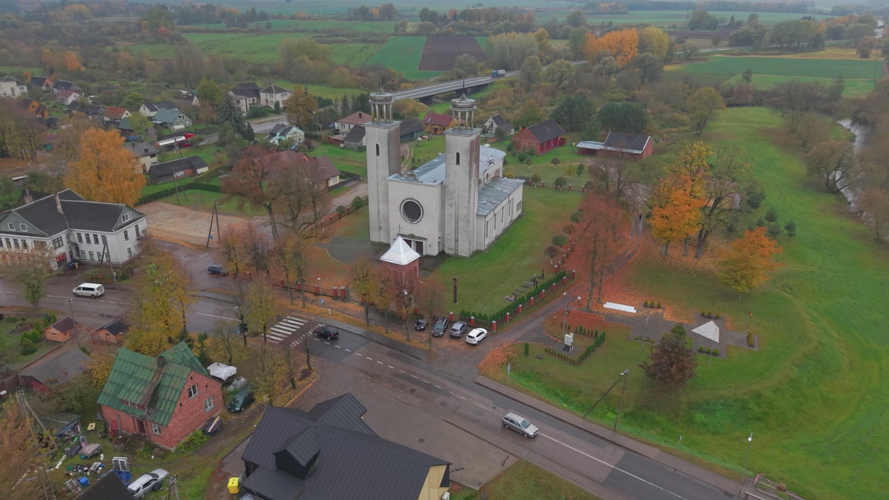 Drone footage captures the Pilviškiai Holy Trinity Church surrounded by autumn foliage in Lithuania. The peaceful village setting, framed by lush fields and serene skies.