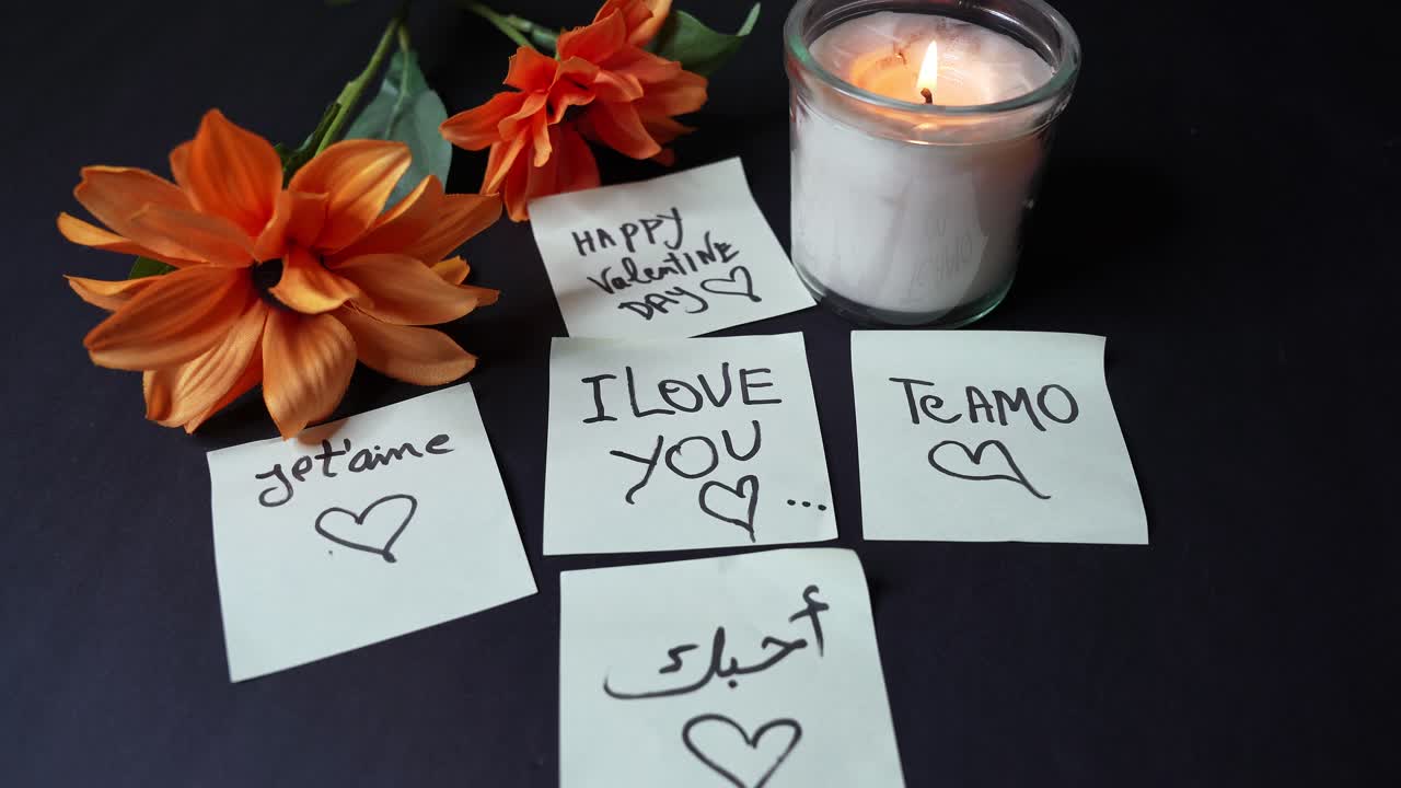 Valentinsdag stilleben med tændt glasfyrfadsstage, orange blomster og håndskrevne post-its med 'Jeg elsker dig' på flere sprog på et mørkt bord, varm afdæmpet belysning