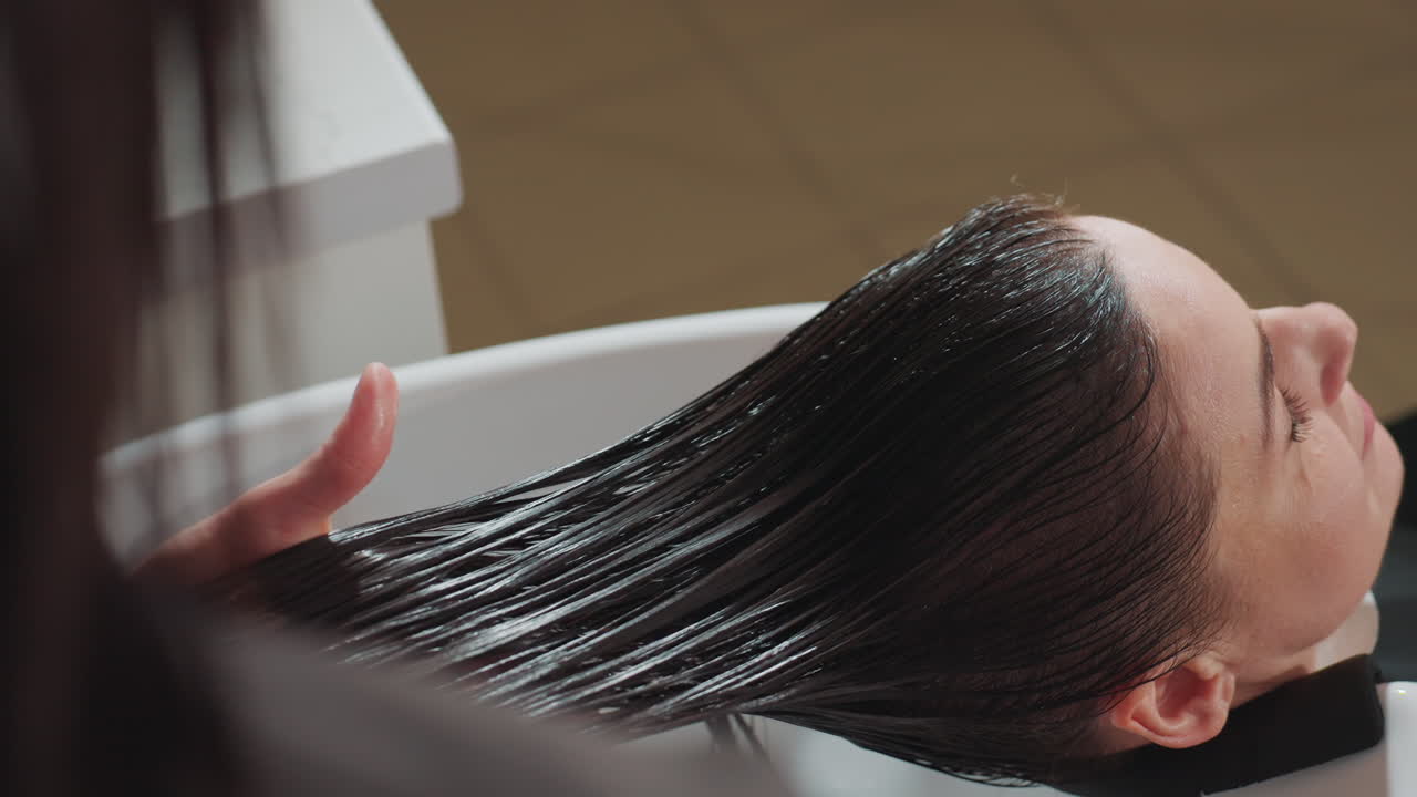 미용실 세면대에서 스타일리스트가 젖은 머리를 빗질하며 편안한 분위기 속에서 부드럽게 헹구고 씻어내세요. 두피를 지지해주고 매끄러운 마무리를 위해 전문적인 클렌징을 제공하며, 물은 천천히 흐릅니다.