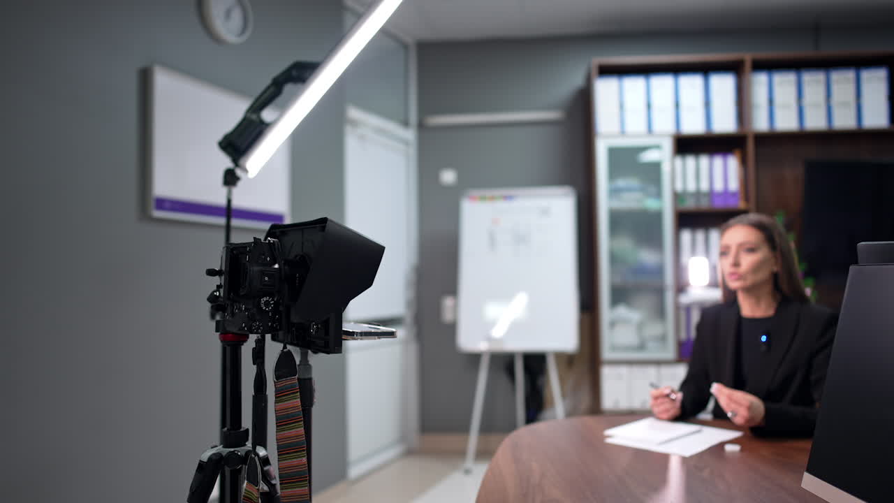 Brunette woman wearing black jacket sitting at desk talks to camera. Influencer speaks passionately creating blog. Blurred backdrop.