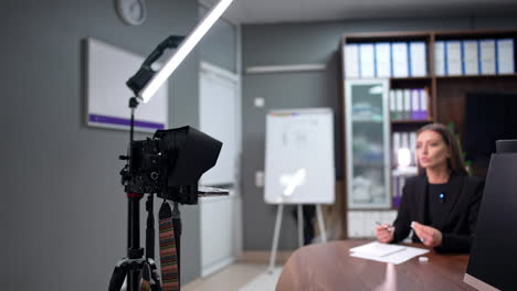 brunette woman wearing black jacket sitting at desk talks to camera. influencer speaks passionately creating blog. blurred backdrop.