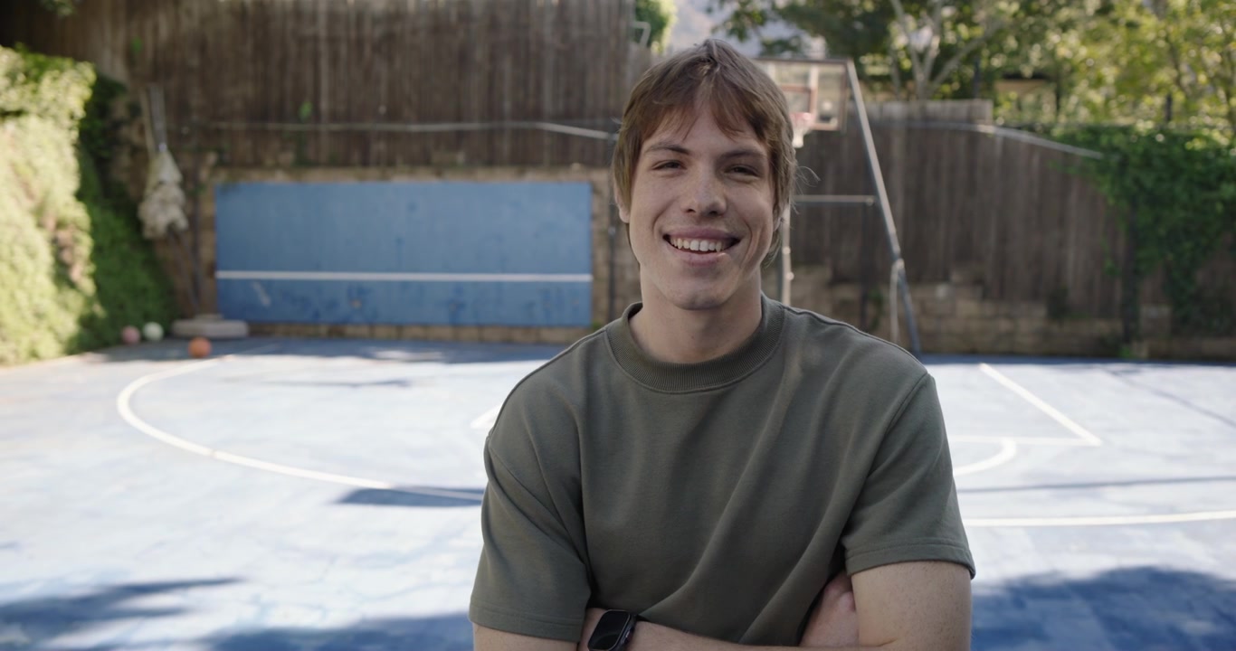 Giovane uomo sui vent'anni in piedi su un campo da basket in giardino con canestro e palloni da basket, la telecamera che zooma, sorridente.