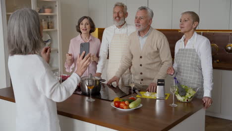 feliz anciana haciendo un selfie con un smartphone en la cocina, sus amigos se ríen y saludan a la cámara