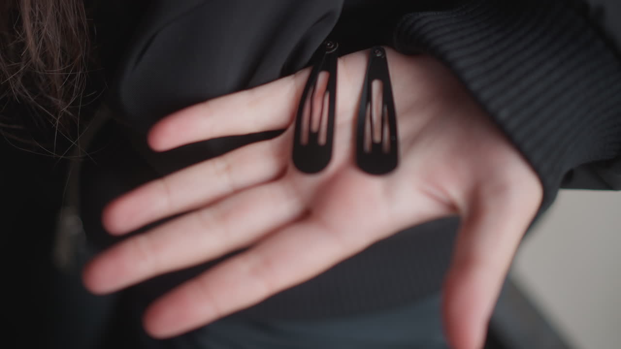 Primo piano della mano di una donna che tiene delle forcine, con la giacca. Atmosfera di strada piovosa e soffusa, manica nera che incornicia il palmo pallido, due fermagli neri appoggiati sui polpastrelli, gesto al rallentatore, sottili riflessi di gocce di pioggia, cinematografico.