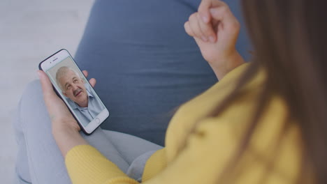 Over-shoulder-view-of-young-woman-daughter-video-calling-old-parent-father-or-mature-friend-using-conference-chat-online-application-on-mobile-phone-screen-at-home-office.-Family-videocall-concept