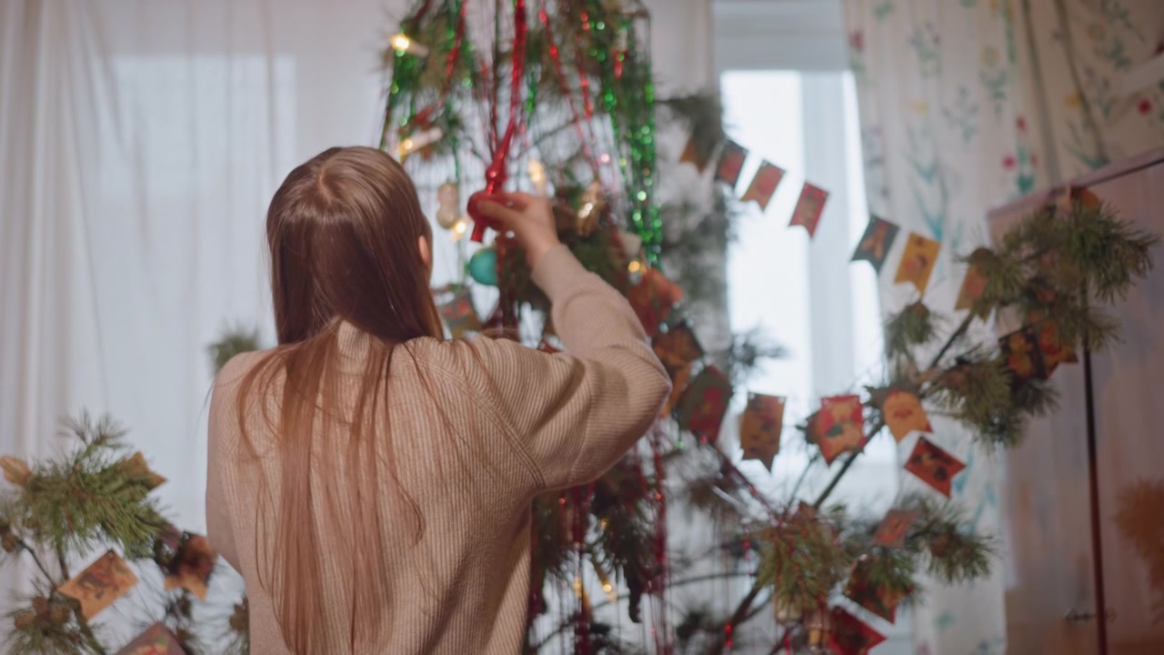 Woman extends to top of tree with decorations. Female individual reaching upper branch adorned with ornaments. Woman with long hair and cozy sweater reaching for decoration atop tall tree
