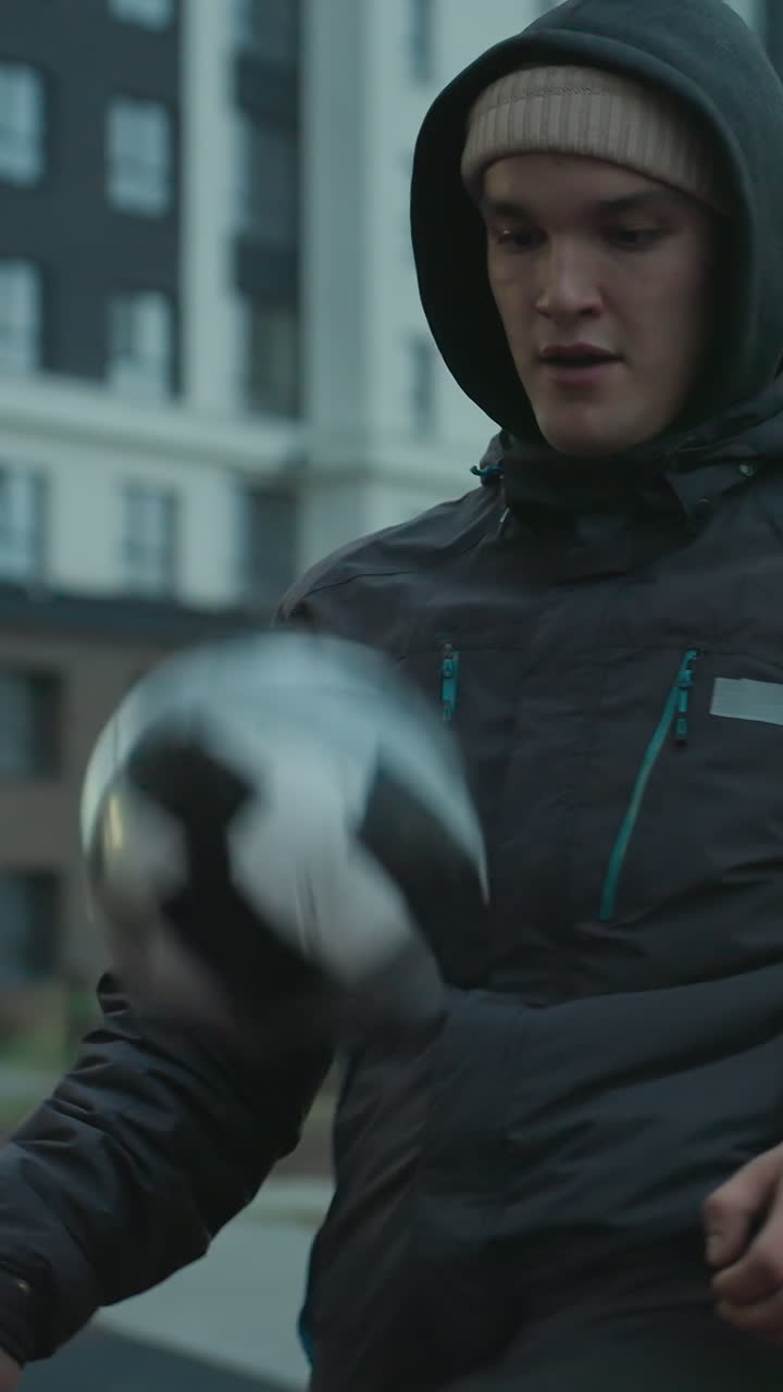 atleta haciendo malabarismos con la pelota con la rodilla durante el entrenamiento en la arena deportiva urbana, fondo con bancos, edificios, equipos de fitness y mujer empujando el portero de bebé en vista borrosa