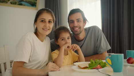 Portrait-of-a-happy-and-joyful-family,-a-brunette-man-in-a-gray-T-shirt,-his-wife,-a-girl-in-a-white-T-shirt,-and-their-little-daughter,-a-brunette-girl-in-a-yellow-dress,-sitting-at-the-family-white-table-and-breakfast-lying-on-plates-in-the-kitchen-in-the-morning