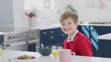portrait of smiling boy wearing school uniform eating breakfast at kitchen counter