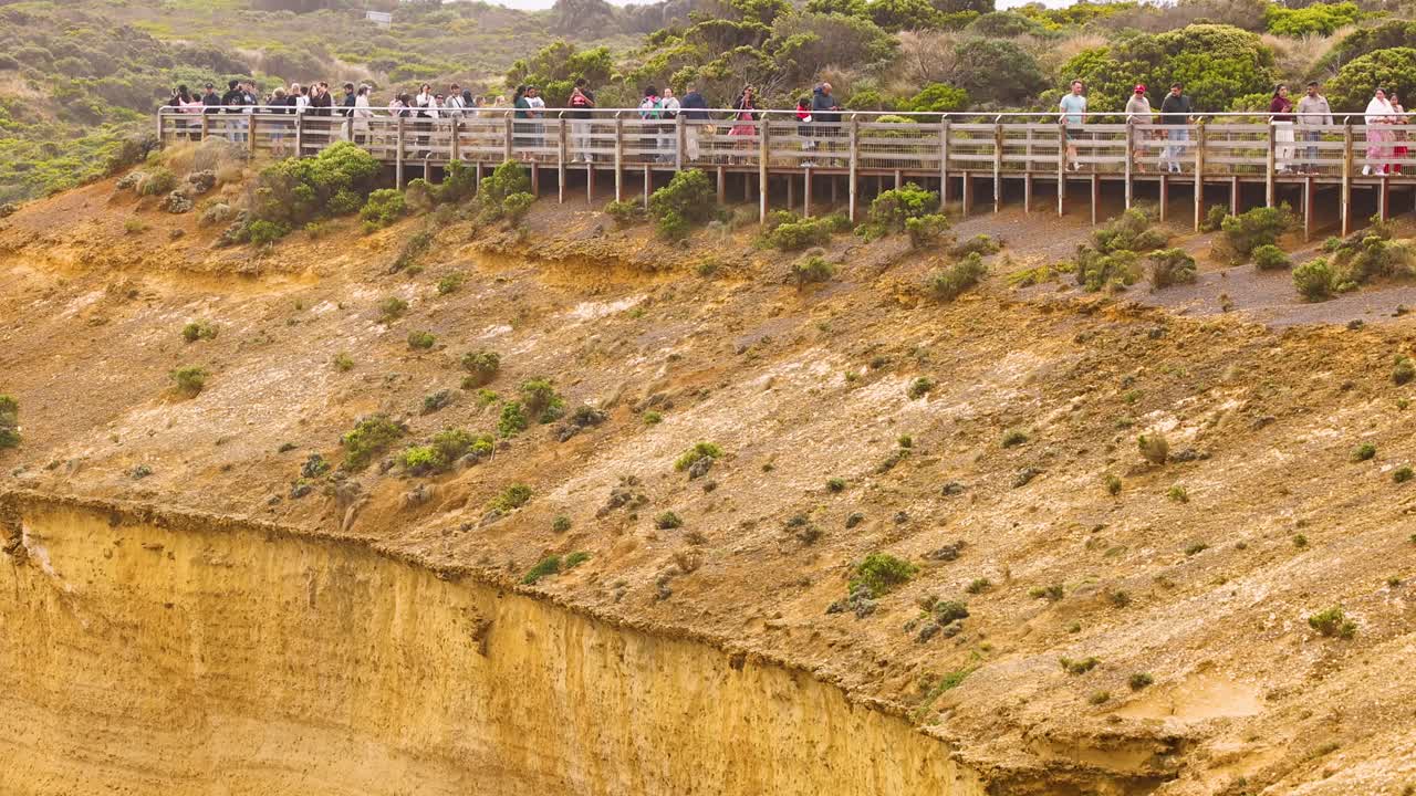 Tourists gather on a viewing platform overlooking rugged cliffs at Port Campbell, Australia. Bright daylight enhances the natural landscape