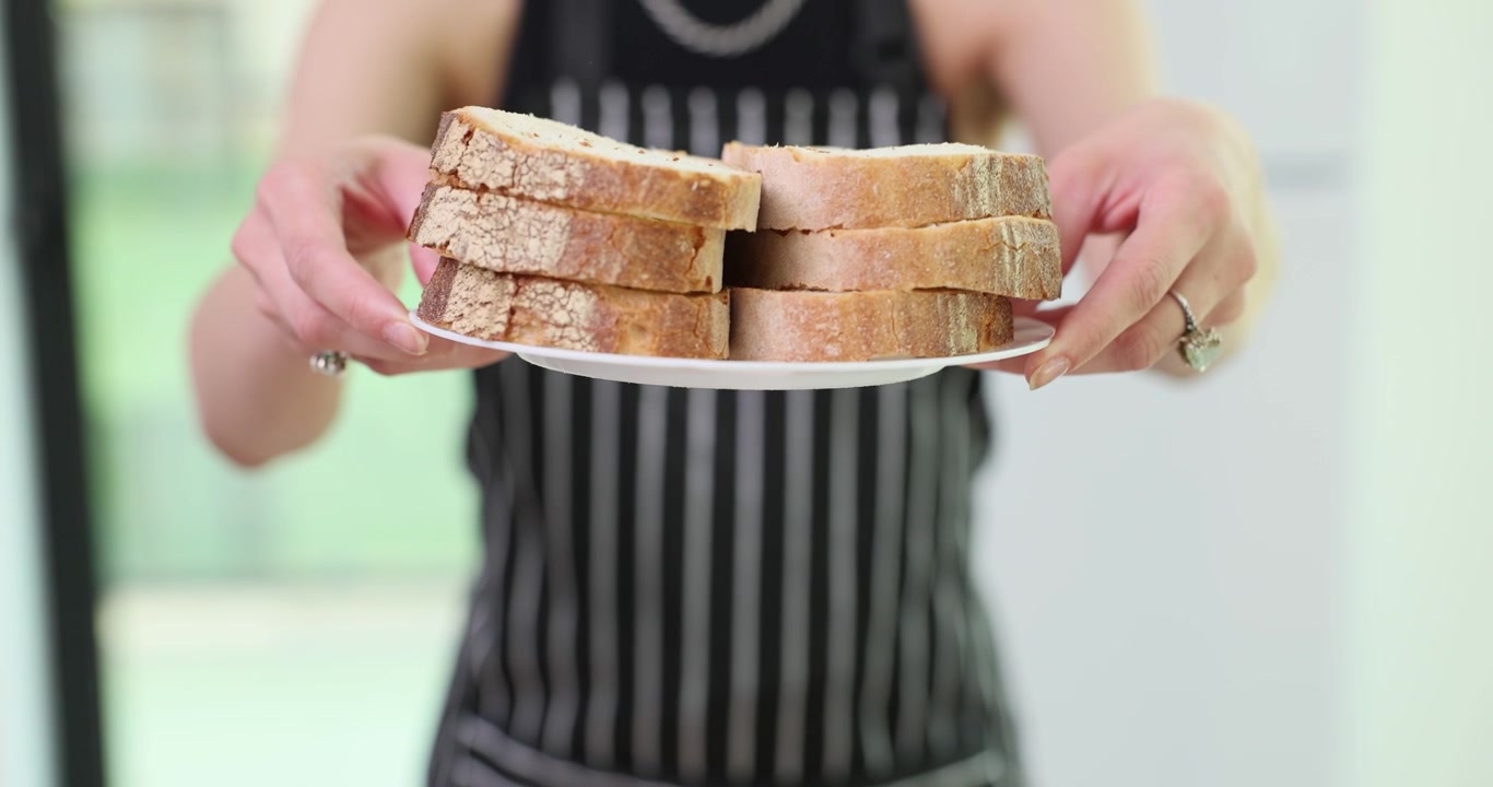 Woman in apron raises plate with bread slices in kitchen