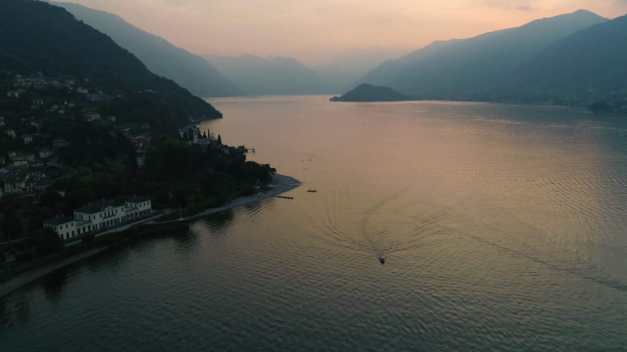 Vista aerea con drone di una piccola barca che attraversa il Lago di Como con la luce dorata del tramonto che si riflette sull'acqua calma, circondato da montagne e da una città lacustre in una leggera foschia.