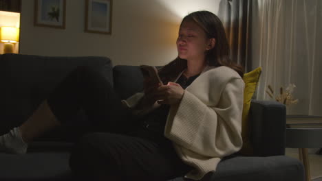 woman spending evening at home sitting on sofa with mobile phone scrolling through internet or social media 7